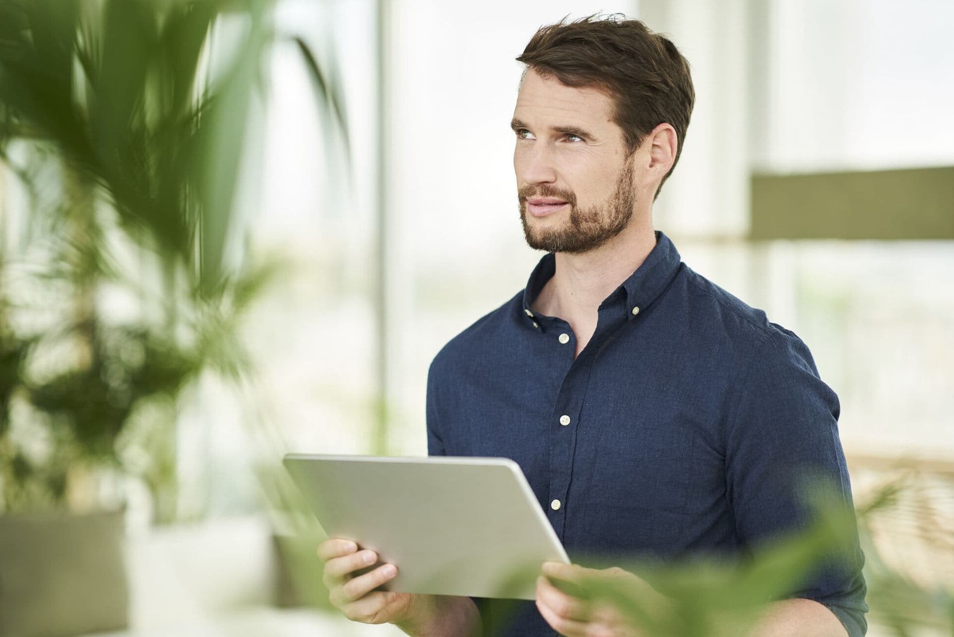 A man sitting barefoot in a bright, modern living room, holding a tablet and gazing out a large window overlooking a sandy, rocky landscape.